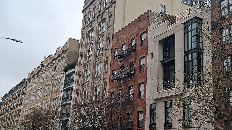 A row of loft buildings of different ages side by side, an ornate limestone building, a plain brick warehouse with a fire escape, and a glass and stone infill building at the end of the block.