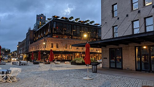 A converted market building at dusk, its brick base carrying a glass walled upper storey with a planted roof deck, outdoor cafe tables and red umbrellas on the Belgian block in front.