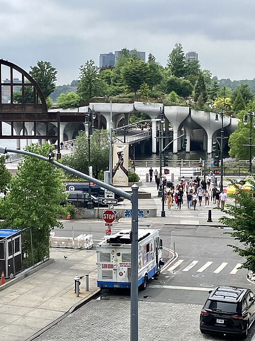 Little Island's raised concrete pillars and planted deck over the Hudson River, a footbridge crossing to it crowded with people out walking.