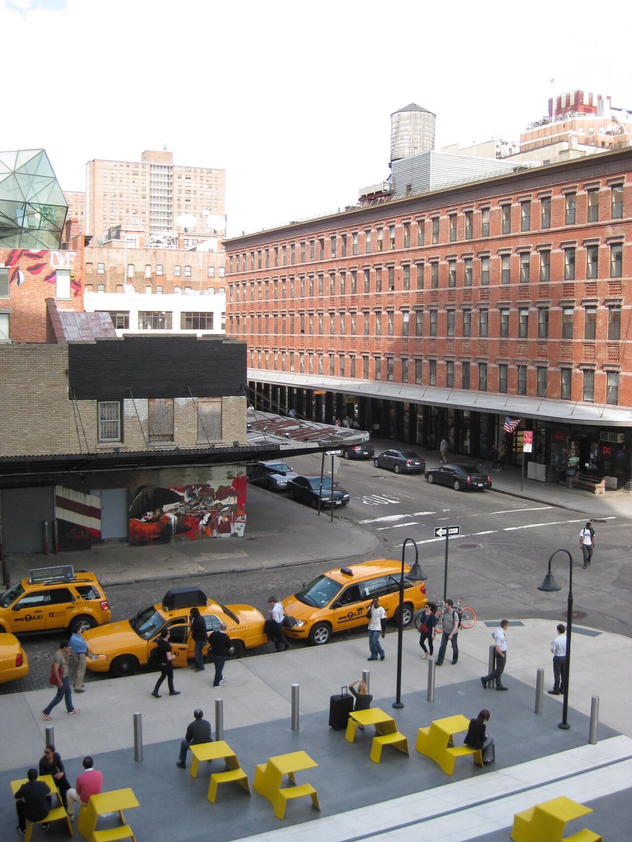 A wide elevated view from the High Line over the 13th Street plaza, a multi storey red brick market building with a metal loading canopy at street level, taxis lined up along the curb below.