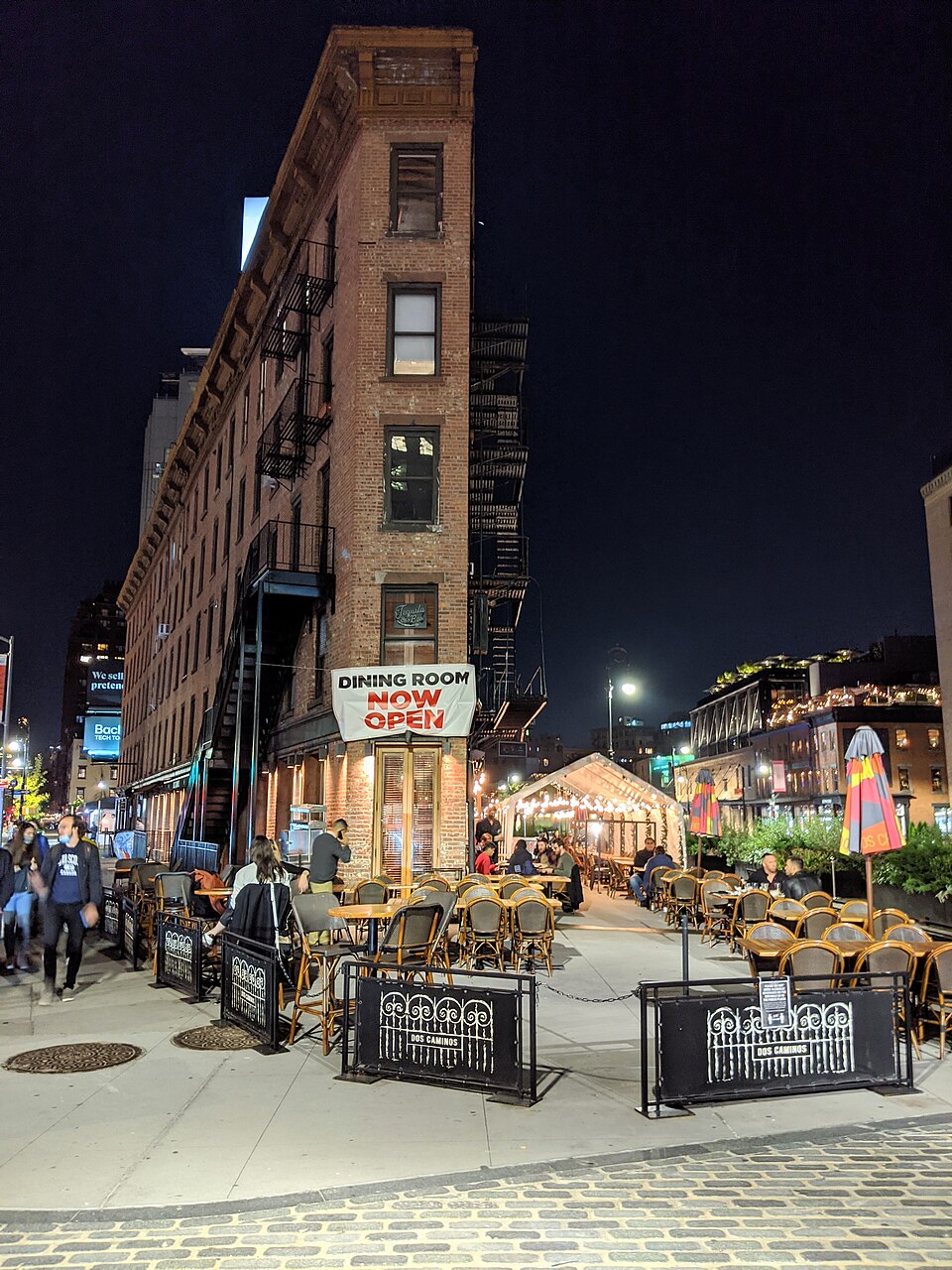 The Triangle Building at night, a narrow six storey brick wedge with a zigzagging fire escape, its ground floor now a restaurant with outdoor tables set out on the Belgian block plaza in front.