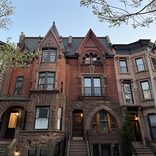Two Queen Anne rowhouses on St John's Place at dusk, red brick over brownstone, with gables, a conical turret and a slate roof.