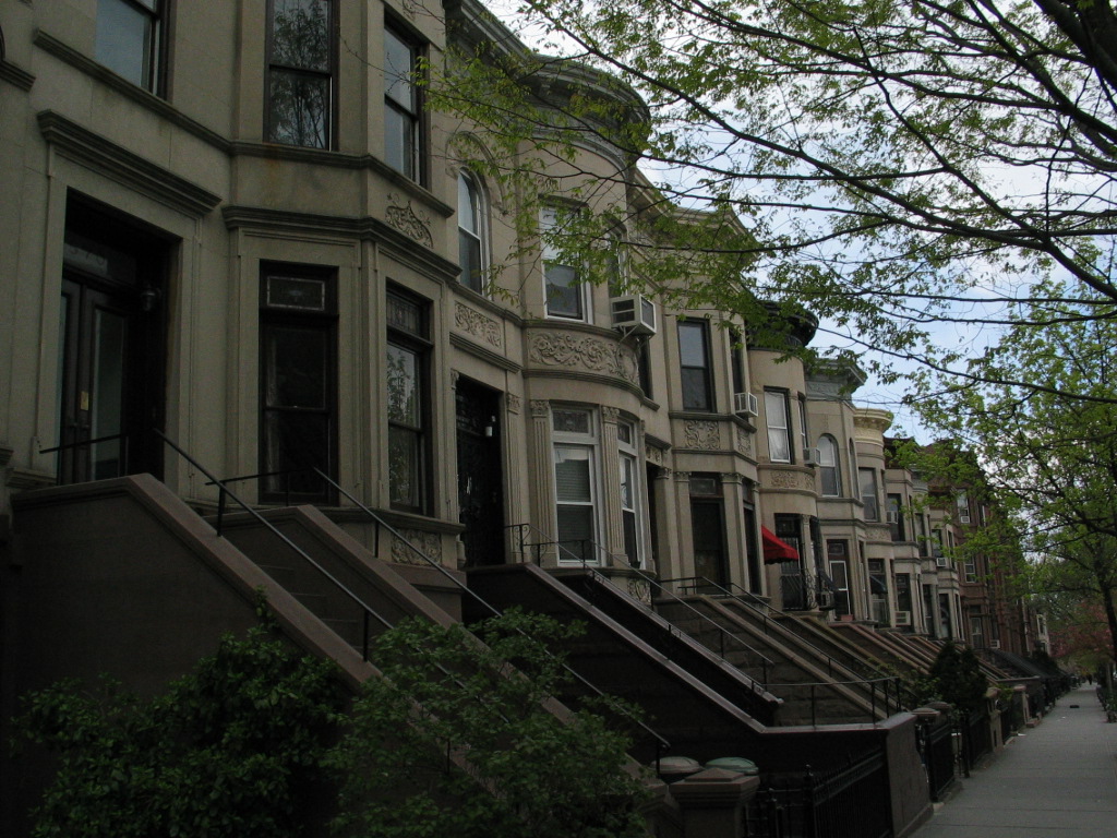 A side street of limestone and brownstone rowhouses seen down the block, with stoops the whole way and street trees over the sidewalk.
