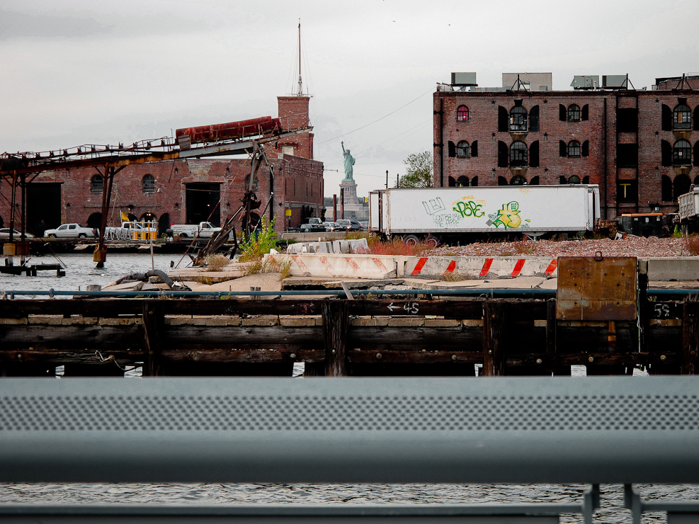 The Statue of Liberty seen between two brick warehouses at the water's edge, a gantry crane over the pier on the left.