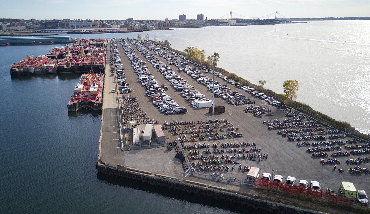 The NYPD impound lot on the pier at Erie Basin, rows of cars and motorcycles with red tank barges moored alongside.
