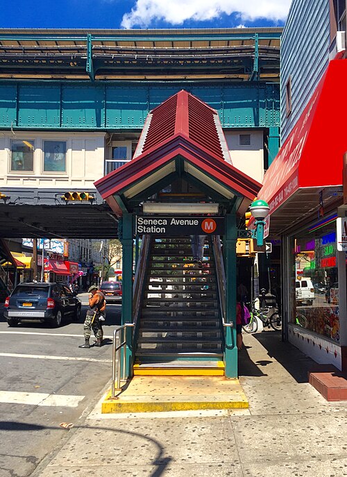 Myrtle Avenue under the el, the Seneca Avenue station stair and storefronts in bright sun.