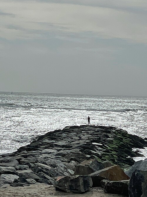 A stone groyne running out into the surf on a grey day, one person standing at the end.