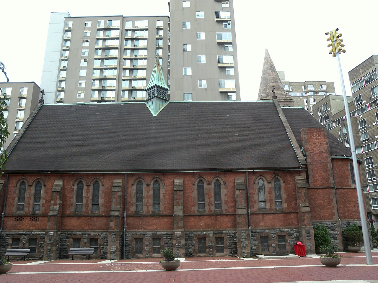 The Chapel of the Good Shepherd in brick and stone with a slate roof and a small copper spire, and apartment slabs standing directly behind it.