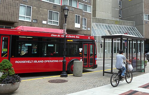 A red island bus at a Main Street stop, with a glass shelter and a concrete apartment block behind it.