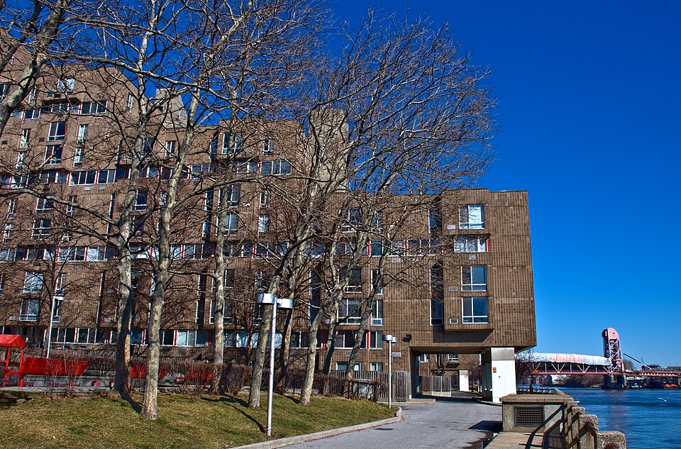 A long brown brick apartment block standing at the water on the island's east side, bare trees in front of it and the red lift bridge from Queens beyond.