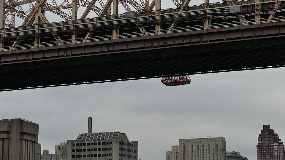 A red tram cabin on the wire, seen past the underside of the Queensboro Bridge with Upper East Side rooftops below it.
