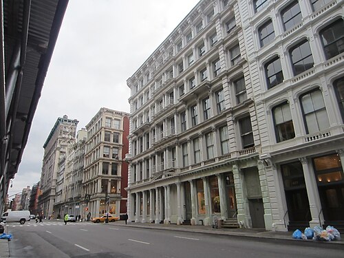 A block of Broome Street on an overcast morning, white painted cast iron on the right with columns and stone steps at street level, a taxi at the far crossing.