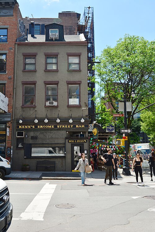 A three floor house at West Broadway and Broome Street with a shingled dormer roof, a painted bar sign across the front and pennants strung over the seating outside.