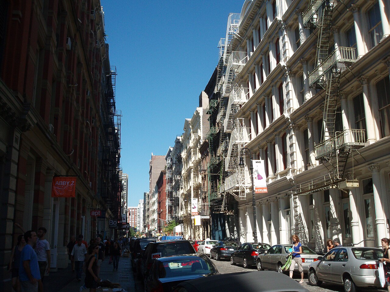 Greene Street looking down the block, cast iron fronts with fluted columns on the sunlit side and long fire escapes on the shaded side, cars parked the whole length of both curbs.