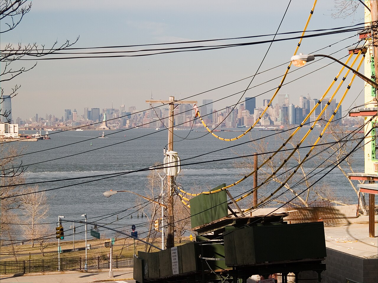 A street on the hill above St. George, overhead wires crossing above it, looking down across the harbor to the Statue of Liberty and the Manhattan skyline.