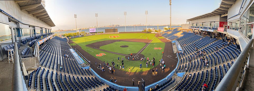 SIUH Community Park's seating bowl and infield from the upper deck, New York Harbor and the Manhattan skyline visible beyond the outfield wall, spectators scattered across the field before a game.