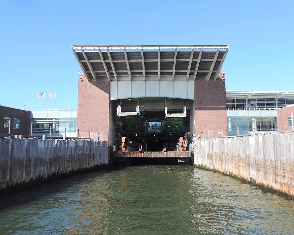 The St. George ferry terminal seen from an incoming boat, an orange ferry easing into its slip under the terminal's canopy on a sunny morning.