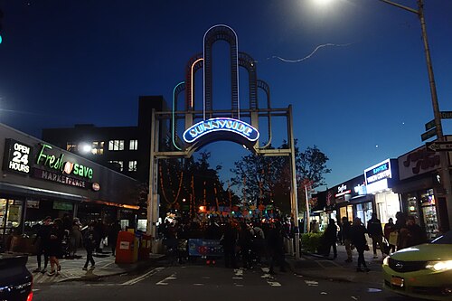 The Sunnyside Arch over 46th Street on the night of a street fair, neon lettering lit and decorations strung down the block.