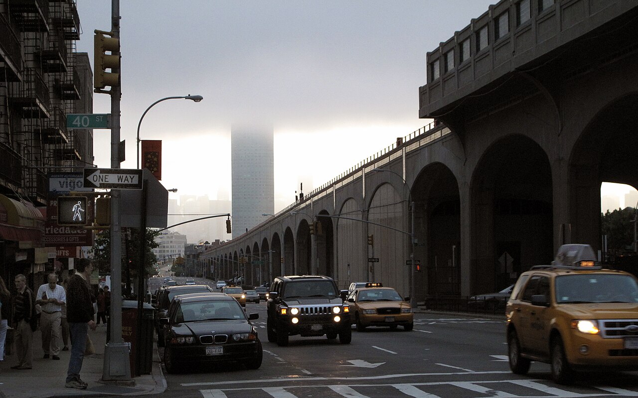 Queens Boulevard at 40th Street after a thunderstorm, taxis in the roadway, the concrete arches of the 7 viaduct along one side and the One Court Square tower in cloud at the far end.