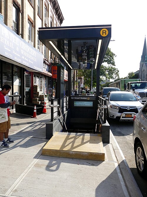 The 53 Street station entrance on Fourth Avenue, a black glass canopy on the sidewalk next to an Indian and Pakistani grocery.