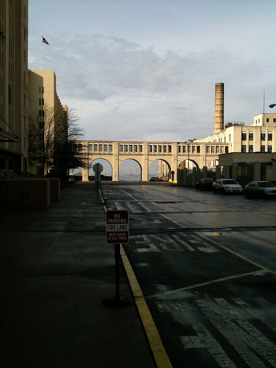 The road inside the Brooklyn Army Terminal, an arcaded bridge across it with the harbor showing through the arches and a brick chimney behind.