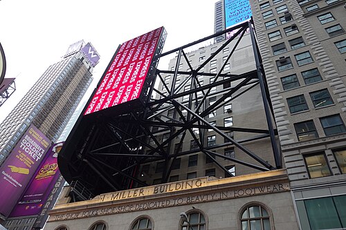 Looking up at the I. Miller Building at Broadway and West 46th Street. THE SHOW FOLKS SHOE SHOP DEDICATED TO BEAUTY IN FOOTWEAR is cut under the cornice, and the steel frame of a screen stands on the roof above it.
