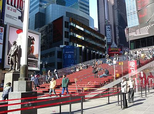 The red glass steps over the TKTS booth in Duffy Square on a bright afternoon, people sitting on them, with the Father Duffy statue at the left and a stock ticker running across the building opposite.