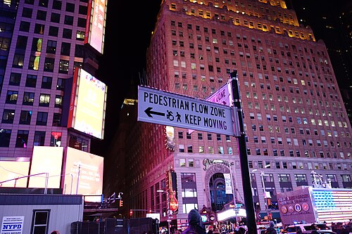 A city sign on the plaza reading PEDESTRIAN FLOW ZONE, KEEP MOVING, with the lit setbacks of the Paramount Building behind it, the Armed Forces recruiting station at the right and an NYPD booth at the lower left.
