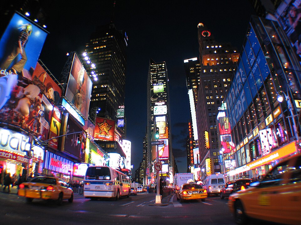 Times Square at night in 2005, seen from the middle of the roadway with taxis and a city bus coming through and lit signs on the buildings either side.