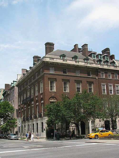 A brick house on a Park Avenue corner, with dormers in the slate roof, tall chimneys above them and a taxi waiting at the crossing.