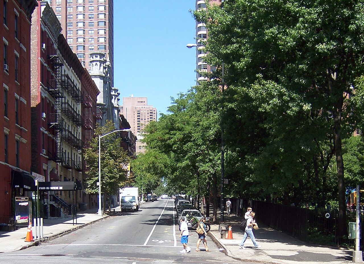 Yorkville at 90th Street and Second Avenue. Tenements with fire escapes stand over the storefronts on the left and there are trees behind a tall fence on the right.