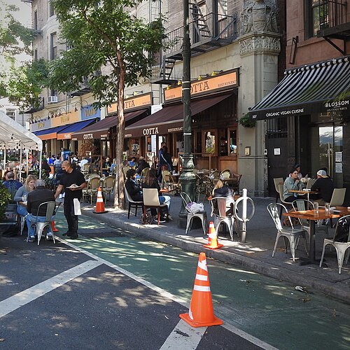 Columbus Avenue near 85th Street, awnings along the storefronts with fire escapes above them and tables out on the sidewalk and in the parking lane.