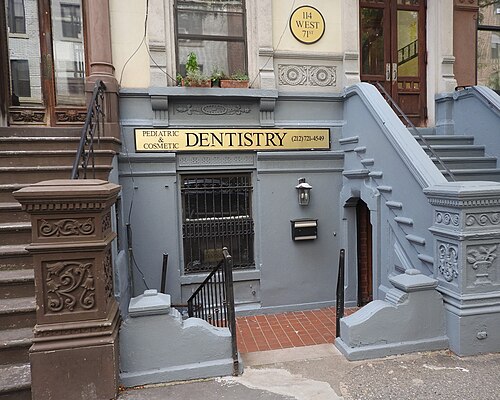 A dentist's office in the garden floor of 114 West 71st Street, under a brownstone stoop painted grey, with a gold sign over the barred window.