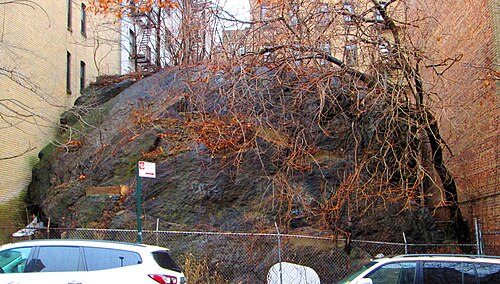 The rock face between 10 and 24 Bennett Avenue, where West 182nd Street would otherwise run, fenced off with cars parked along it.