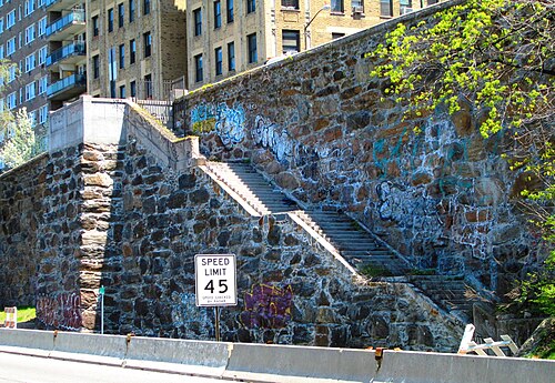Two flights of stone steps down a rubble retaining wall from Plaza Lafayette to the Henry Hudson Parkway, with apartment buildings along the top.
