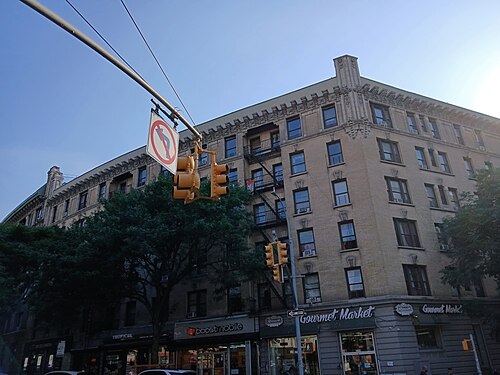 The corner of Edward M. Morgan Place in the Audubon Park Historic District, six stories of tan brick with fire escapes and a grocery and a phone shop underneath.