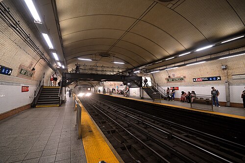 A 1 train coming into the 168th Street station, headlights on, under the tiled barrel vault with the iron footbridge across it.