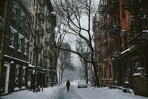 A West Village street in heavy snow, five story brick buildings with fire escapes on both sides and one person walking down the middle of the roadway.