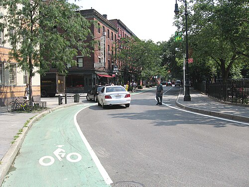 Bleecker Street looking south from Eighth Avenue, low brick buildings with awnings over the shopfronts and a green bike lane along the near curb.