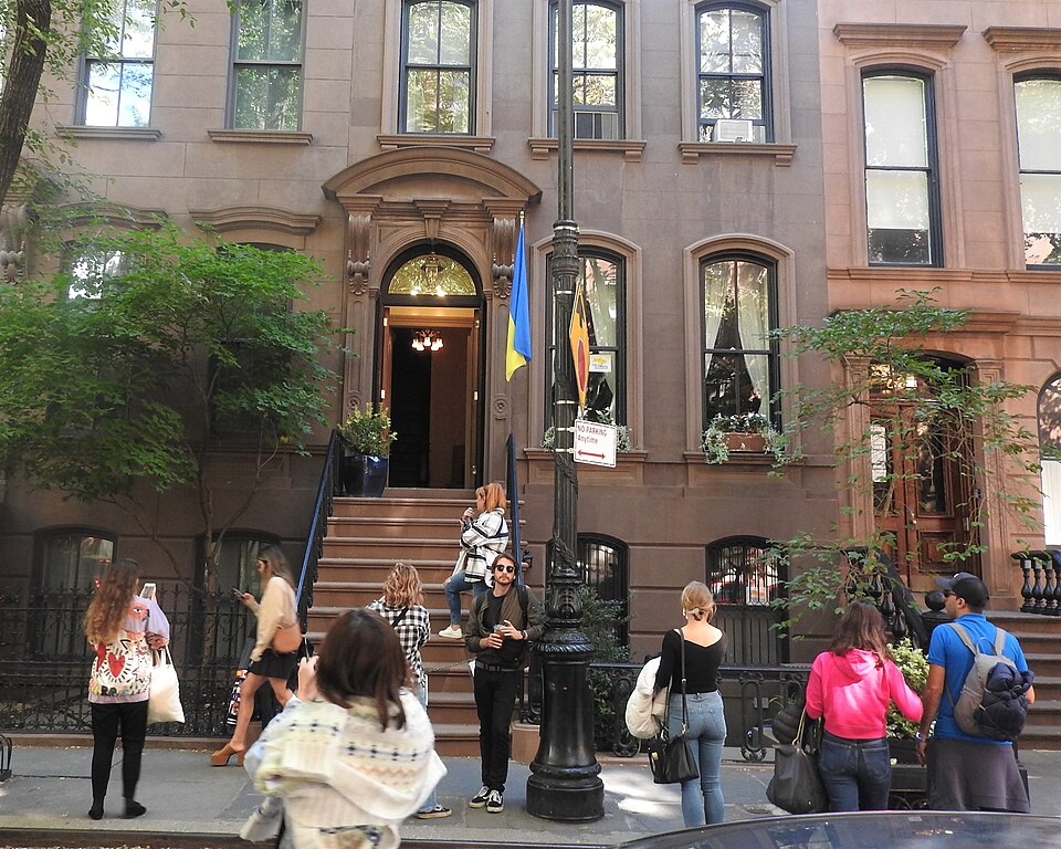 66 Perry Street on a cloudy afternoon, a brownstone stoop under a round arched doorway, with about a dozen people standing on the sidewalk in front of it taking photographs.