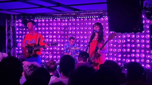 The stage at Baby's All Right on Broadway, three musicians against a wall of round bulbs lit purple, the audience in silhouette across the bottom of the frame.