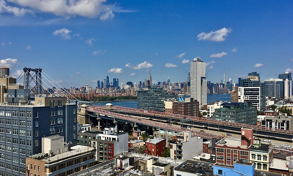 The blocks at the Brooklyn foot of the Williamsburg Bridge from the top of 424 Bedford Avenue, the red approach spans running left to right over low roofs and water tanks, Midtown across the river.