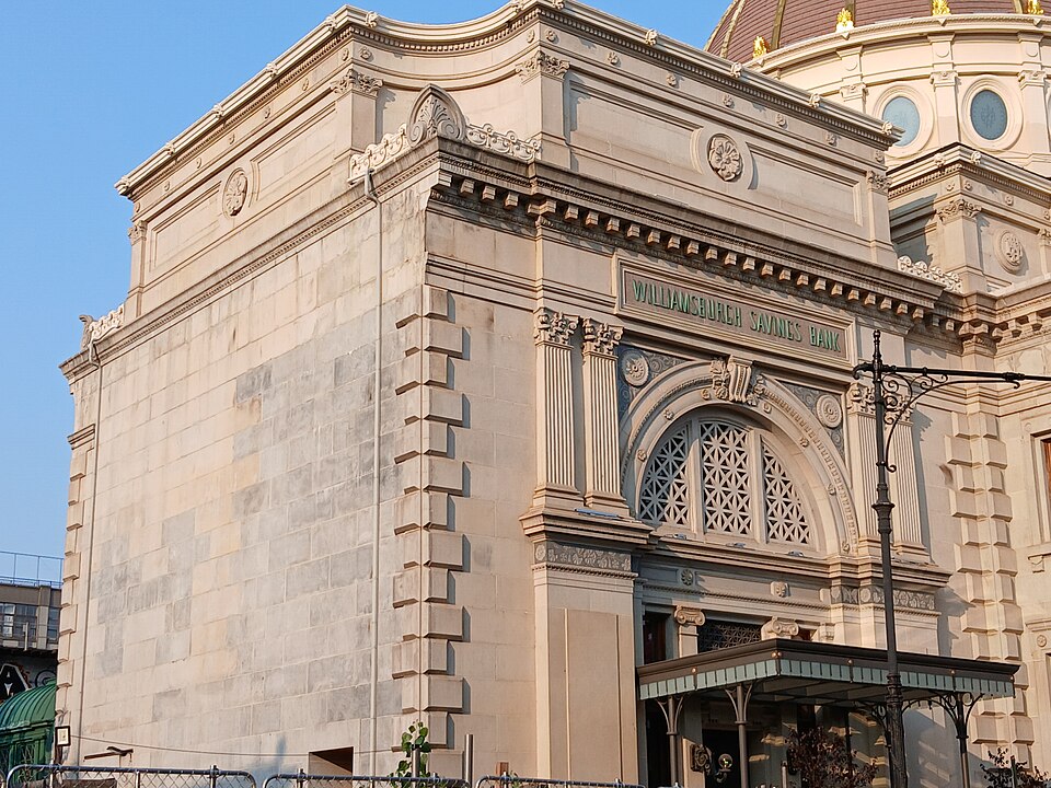 The corner of the Williamsburgh Savings Bank at Broadway and Driggs Avenue in evening light, the name cut into the stone above an arched grille, the dome behind it.
