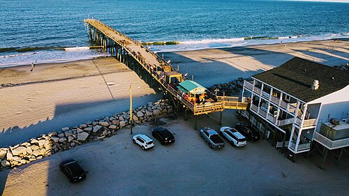 An aerial view of the wooden fishing pier on pilings over the water, a two storey pier house and parking lot at its foot.