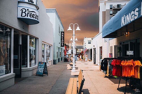 A boardwalk corridor between a clothing shop and an arcade, string lights and lamp posts down the centre, apparel racks set out front.