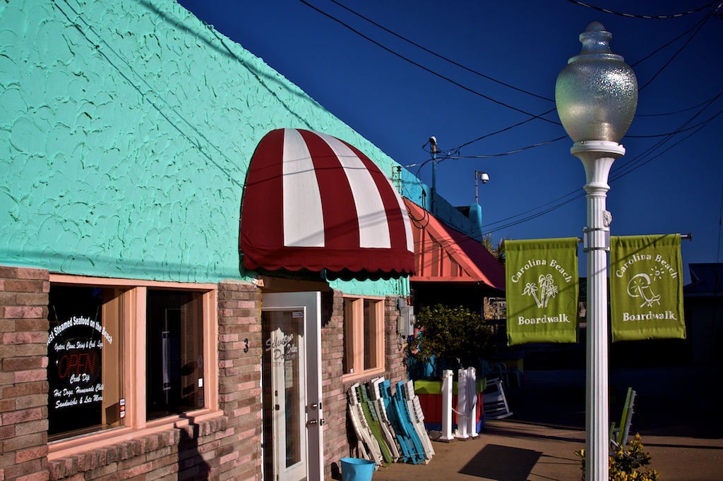 A turquoise stucco storefront advertising steamed seafood, a red and white striped awning over the door, a Carolina Beach Boardwalk banner hanging from the corner lamp post.
