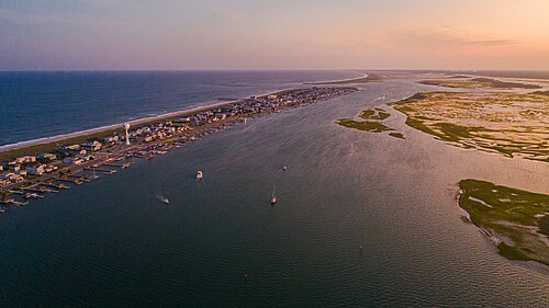 An aerial sunset view of the barrier island's south end, beach houses and docks on the sand strip with open sound and salt marsh behind it, sailboats moored in the channel.