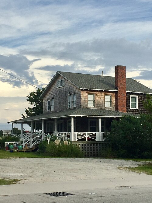 A two storey cedar shingle beach cottage with a wide wraparound porch, white railings, a red brick chimney, and pampas grass by the front steps.
