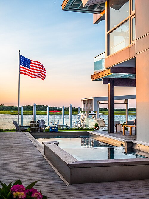 A contemporary glass fronted waterfront house with a rooftop deck, a small infinity edge pool, and a boat dock on the sound behind it.