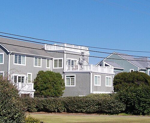 A row of grey shingle beach houses with white trim, roof decks, an outside spiral stair, and low hedges along the front walk.
